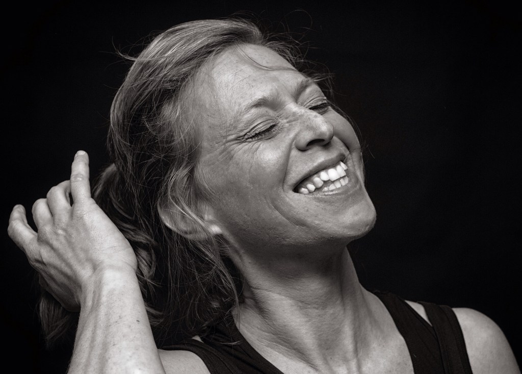Black and white candid portrait of a smiling woman with eyes closed, brushing her hair back in a moment of pure joy.