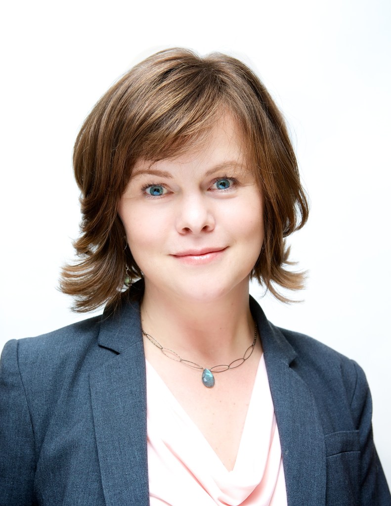 Professional woman with short brown hair, wearing a blazer and necklace, smiling in front of a white background