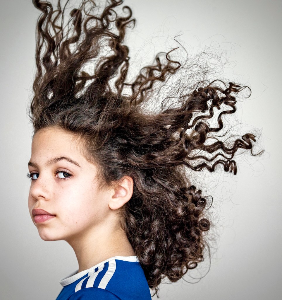 Young girl in a blue jersey with long curly hair blown outward, captured in profile with a confident expression