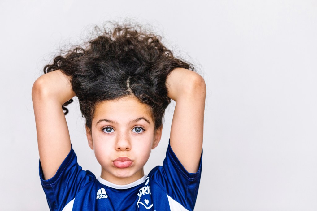Young girl with curly hair raised and blue soccer jersey, looking directly at the camera with a playful expression.
