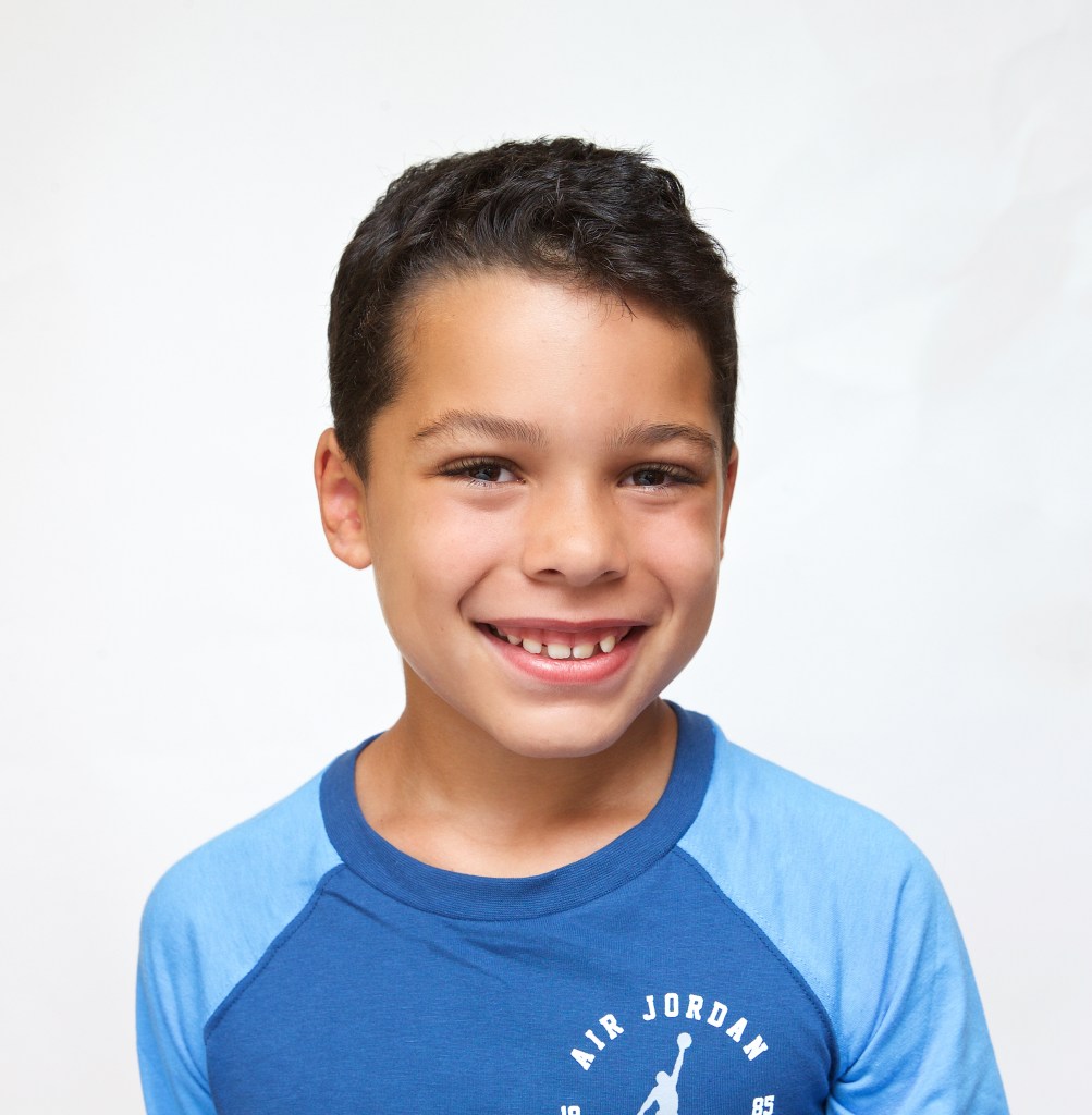 Smiling young boy with short dark hair, wearing a blue Air Jordan shirt, photographed against a white background