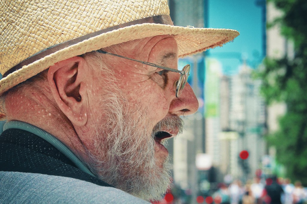 Side profile of an elderly man with a straw hat and glasses, smiling against a blurred city background.