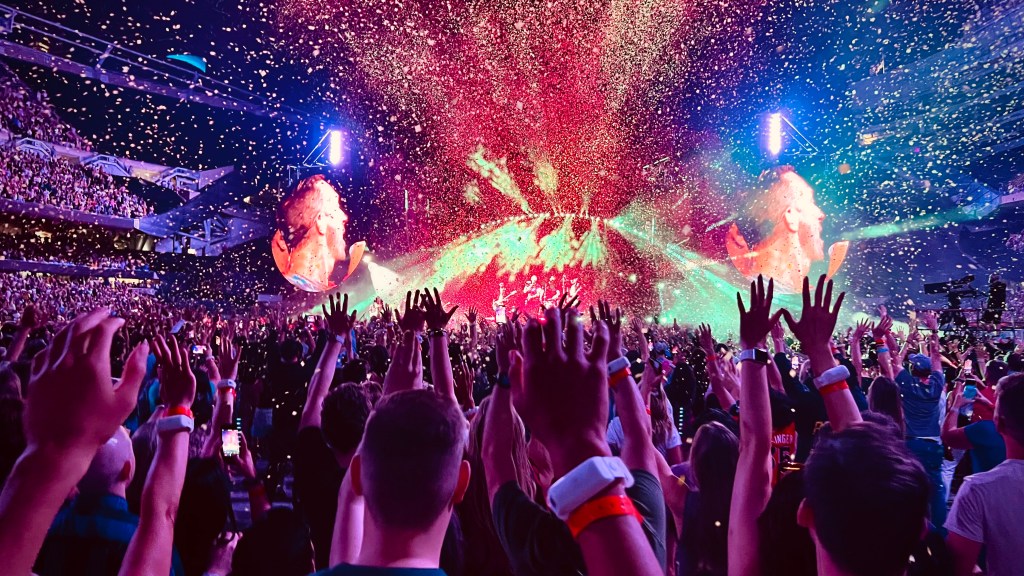 A vibrant concert scene with a lively audience raising their hands, colorful confetti falling, and bright stage lights illuminating the performers in the background.