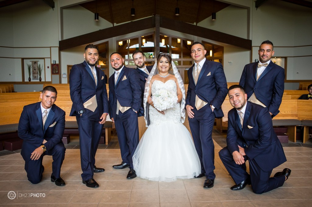 A bride in a white wedding dress holding a bouquet stands in the center with six groomsmen around her, all wearing navy blue suits and cream ties, posing in a chapel setting.