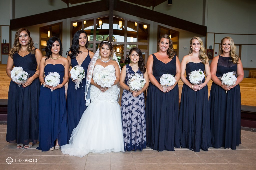 A group of bridesmaids in navy blue dresses, smiling together with the bride in a white gown, holding bouquets of white flowers, standing in an indoor setting.