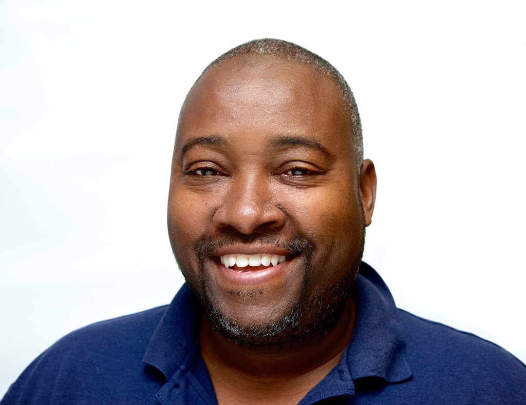 Headshot of a smiling man with short hair wearing a navy blue polo shirt, set against a white background.