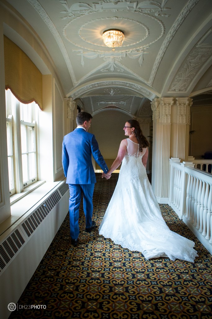 A bride and groom walking hand in hand down a well-lit corridor decorated with intricate architectural details and a patterned floor.