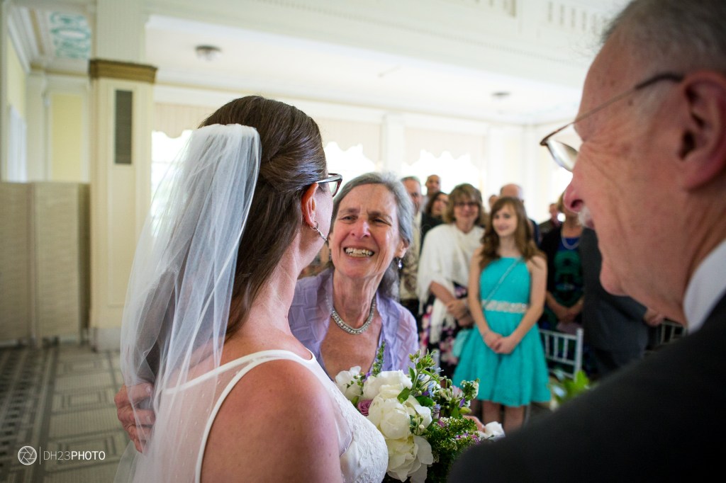 A bride in a white dress and veil is smiling while being greeted by an older woman, likely a family member, in a wedding setting. Guests are seated in the background, adding to the celebratory atmosphere.