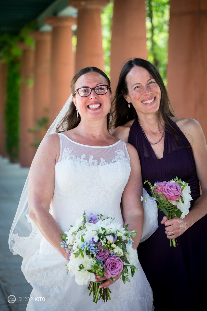 Two women standing together, one in a white wedding dress and veil holding a bouquet of flowers, and the other in a purple dress holding a smaller bouquet. They are smiling and posed in a sunny outdoor setting with columns in the background.