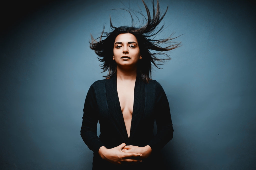 A woman with flowing hair stands confidently against a dark blue background, wearing a deep V-neck top and with her hands clasped in front of her.