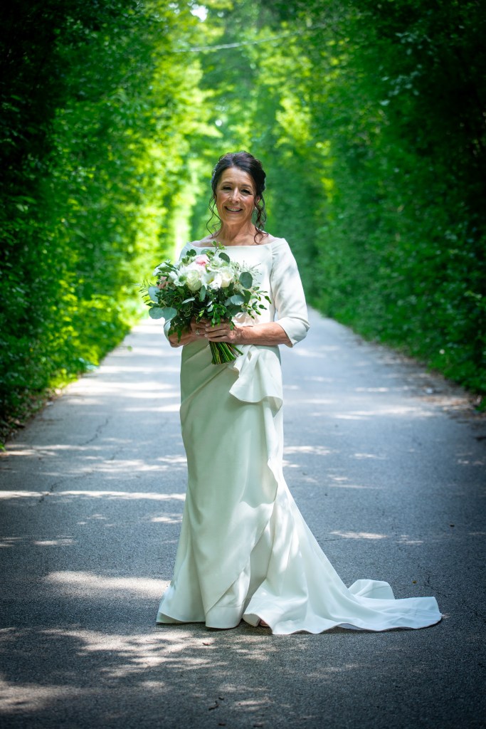 A smiling woman in a white wedding dress holds a bouquet of flowers while standing on a tree-lined path.