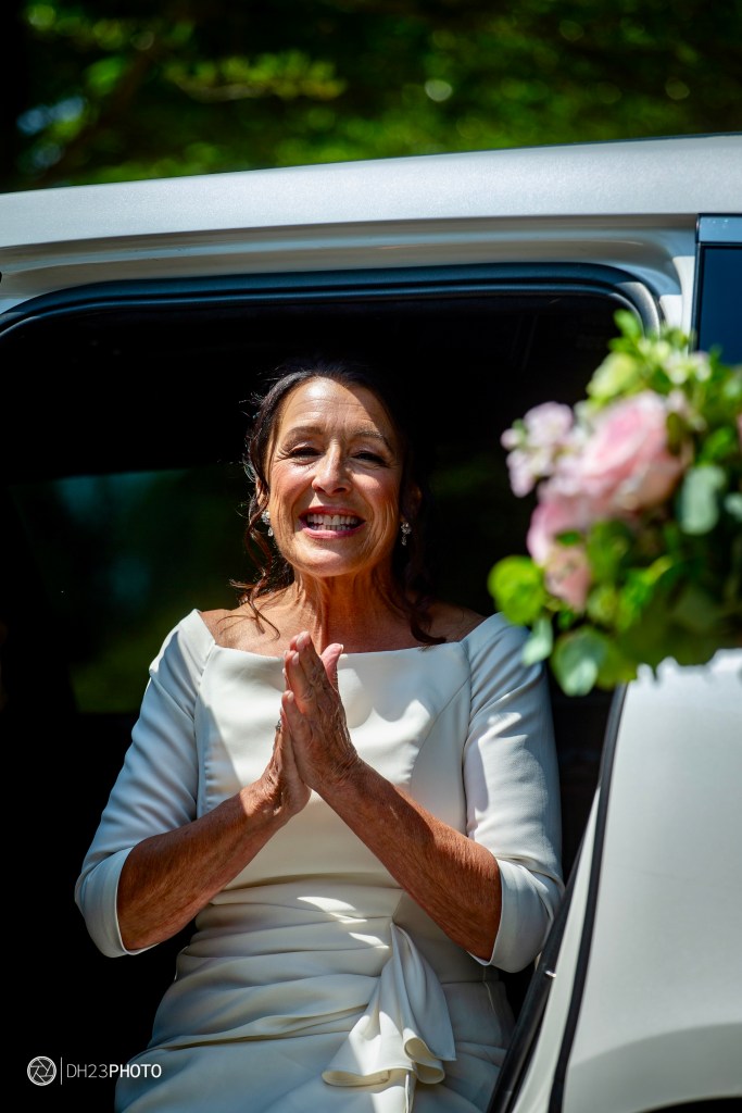 A smiling woman in a white dress is sitting in a car, holding her hands together in a gesture of gratitude or joy, with a bouquet of flowers visible nearby.