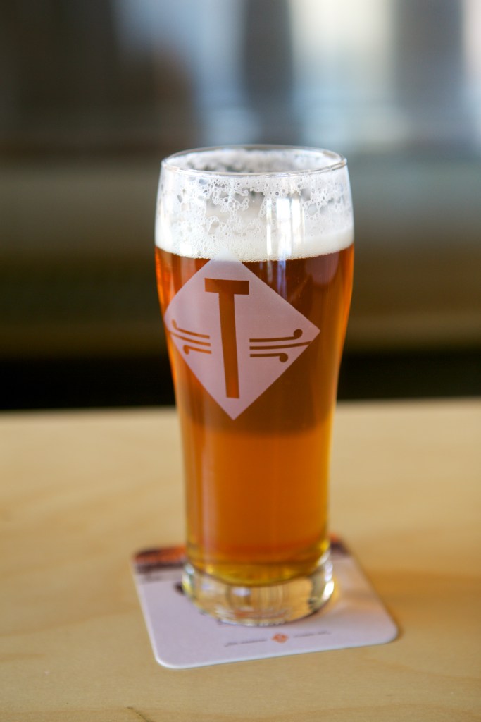 A pint of beer in a clear glass with a logo on it, placed on a coaster on a wooden table.