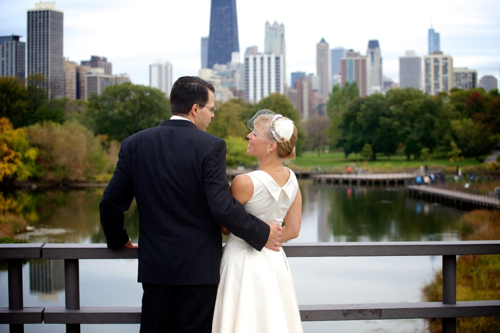 A couple stands on a bridge overlooking the Chicago skyline and a serene body of water. The woman wears a wedding dress and holds her hair back with a decorative veil, while the man is dressed in a formal suit. They are embracing and looking at each other with smiles.