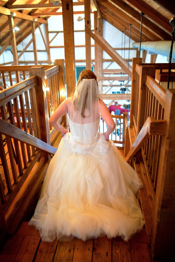 A bride in a flowing white wedding dress descends wooden stairs in a rustic venue, with beams and warm lighting around her.