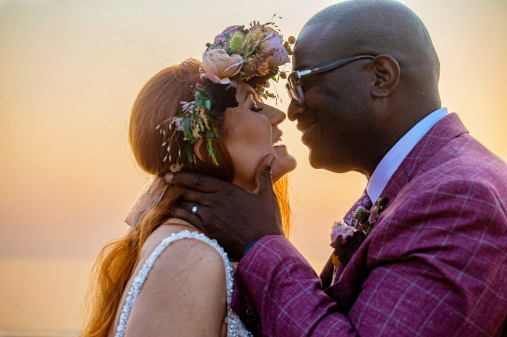 A close-up of a bride and groom sharing an affectionate kiss at sunset, with the bride wearing a floral crown and the groom in a patterned suit.
