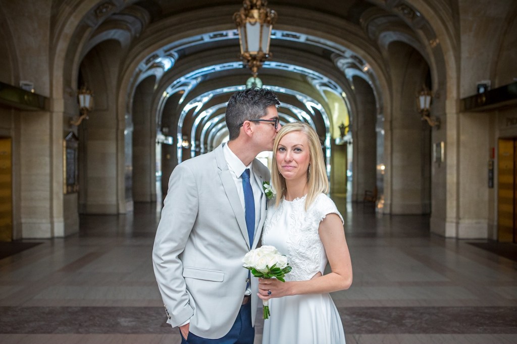A couple posing in an elegant interior, with the man gently kissing the woman's cheek. The woman holds a bouquet of white roses, both dressed in formal attire.