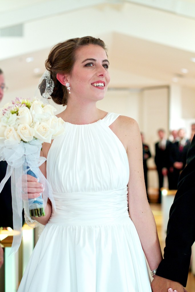 A bride in a white wedding dress holds a bouquet of white roses, smiling while standing next to her partner.