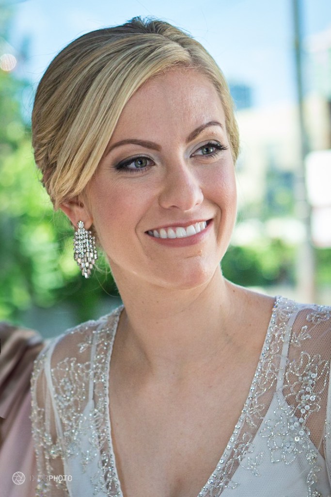 A woman with long blond hair and elegant earrings, smiling while wearing a beautifully detailed, light-colored dress with embellishments, in a bright outdoor setting.