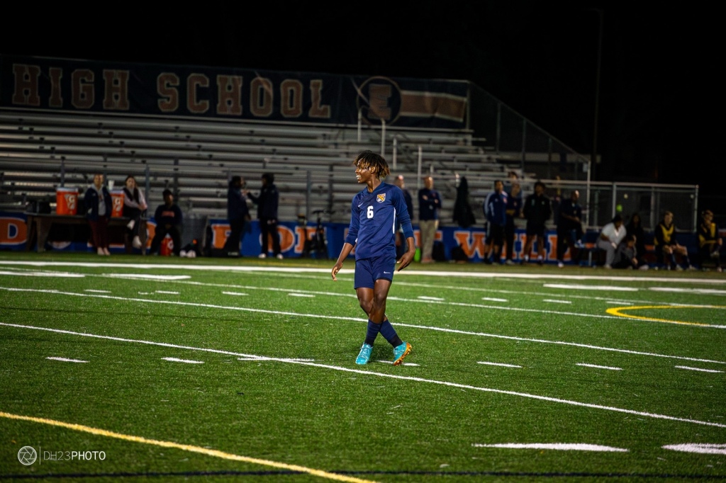 A high school soccer player wearing a blue uniform and number 6 is walking on the field at night, with spectators in the background.