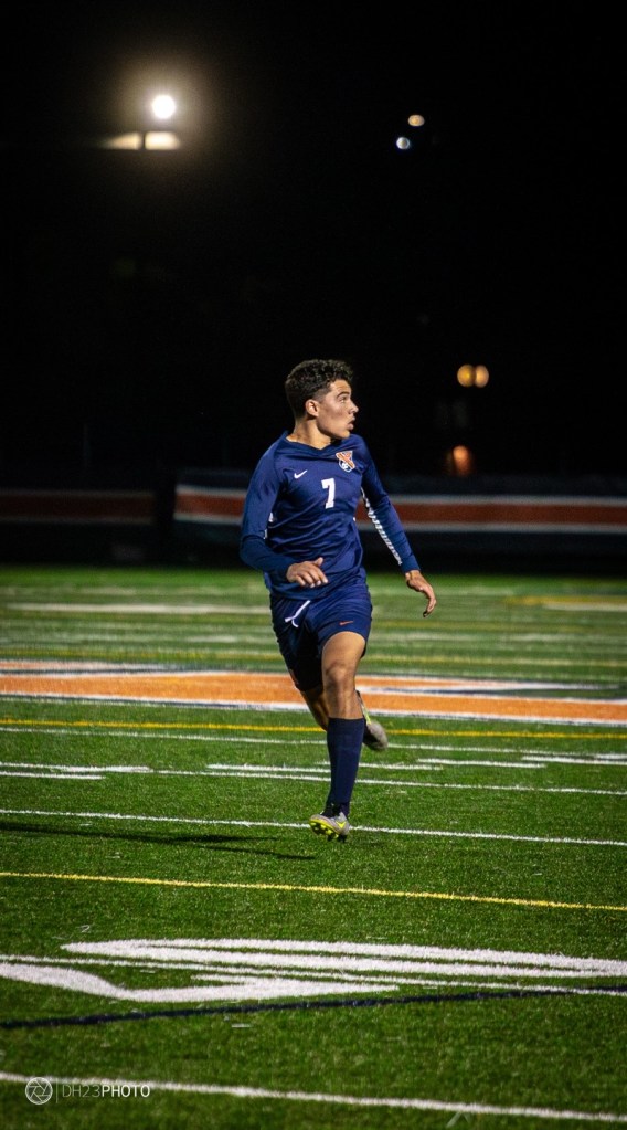 A young soccer player wearing a blue jersey with the number 7 is running on a well-lit field at night.