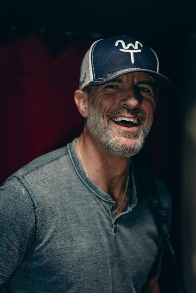 A smiling man with a beard wearing a gray shirt and a cap, standing in front of deep red curtains.