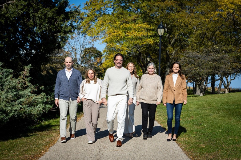 A group of six real estate professionals walking together on a pathway surrounded by greenery and trees during a sunny day.